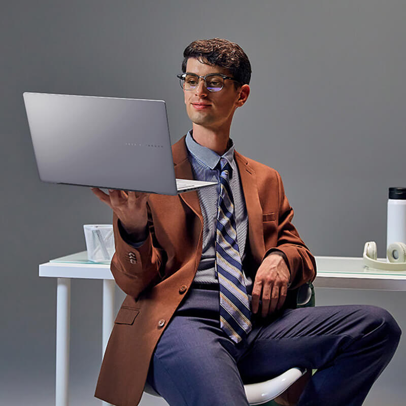 Ein junger Mann in einem formellen Outfit sitzt vor einem Schreibtisch und balanciert einen ASUS Vivobook-Laptop in seiner rechten Hand