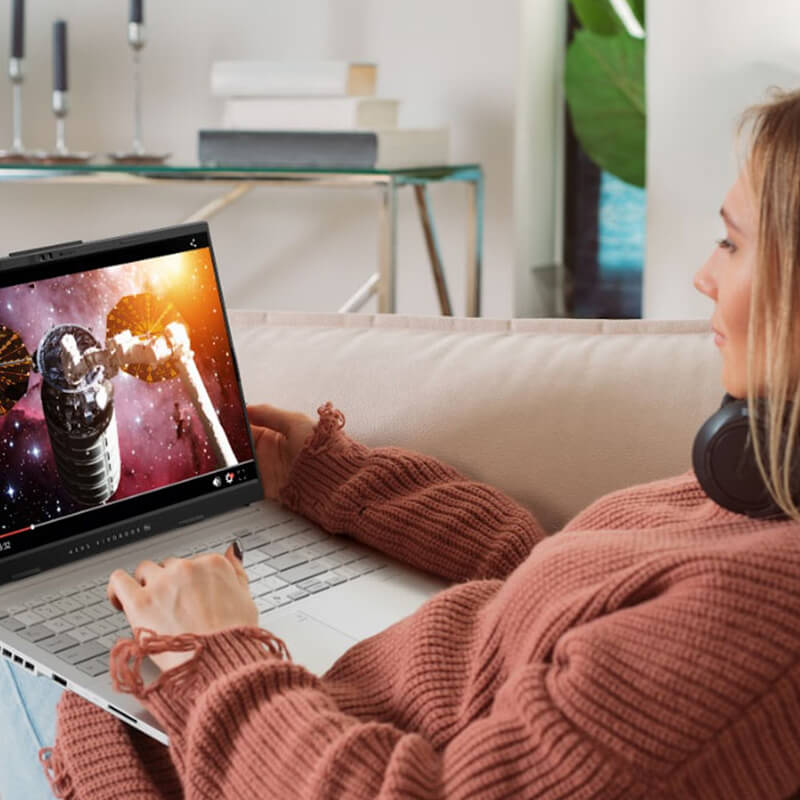 Eine junge Frau sitzt auf einem rosa Sofa mit einem ASUS-Laptop auf dem Schoß und sieht sich ein Online-Video an.